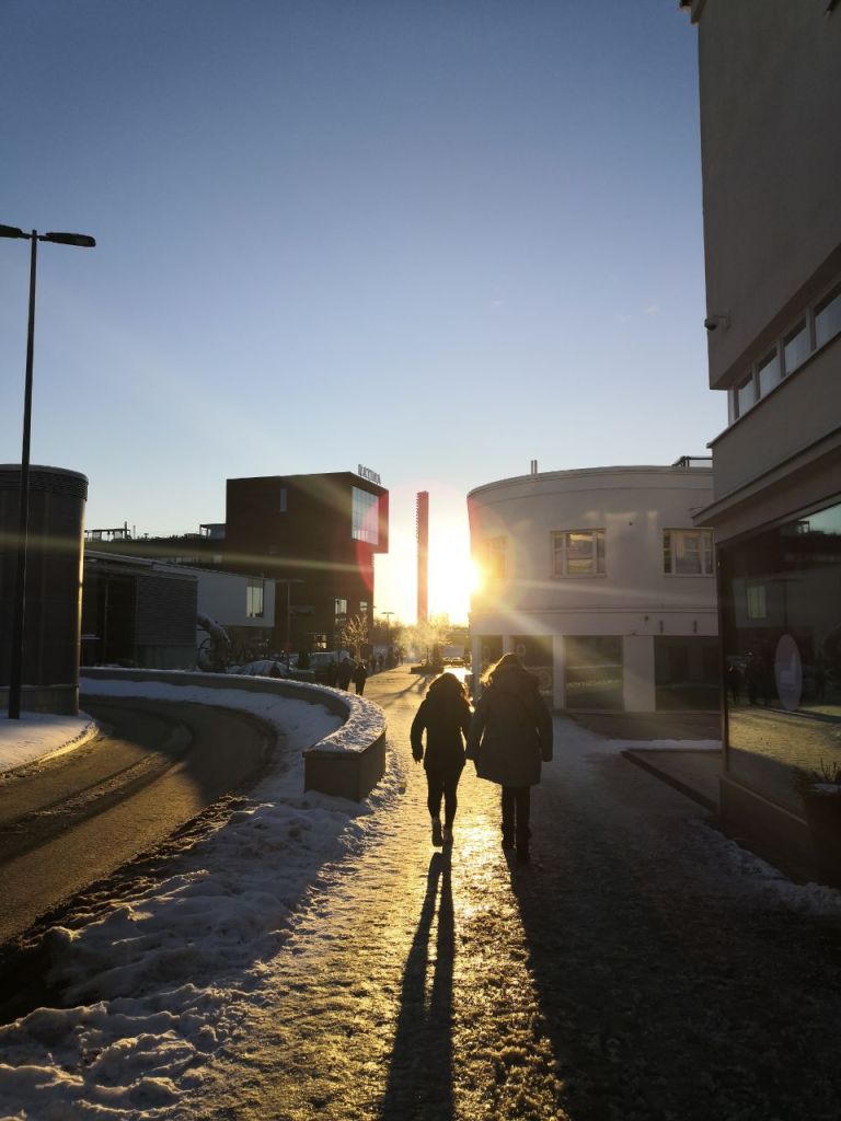 Kaupunkimaiseman jalkakäytävä, jolla kävelee kaksi henkilöä kohti auringonlaskua